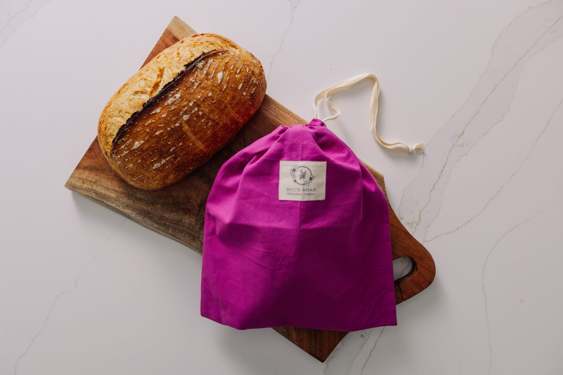 Loaf of bread on a wooden board with a Meadow Magic drawstring bag next to it on a white surface.