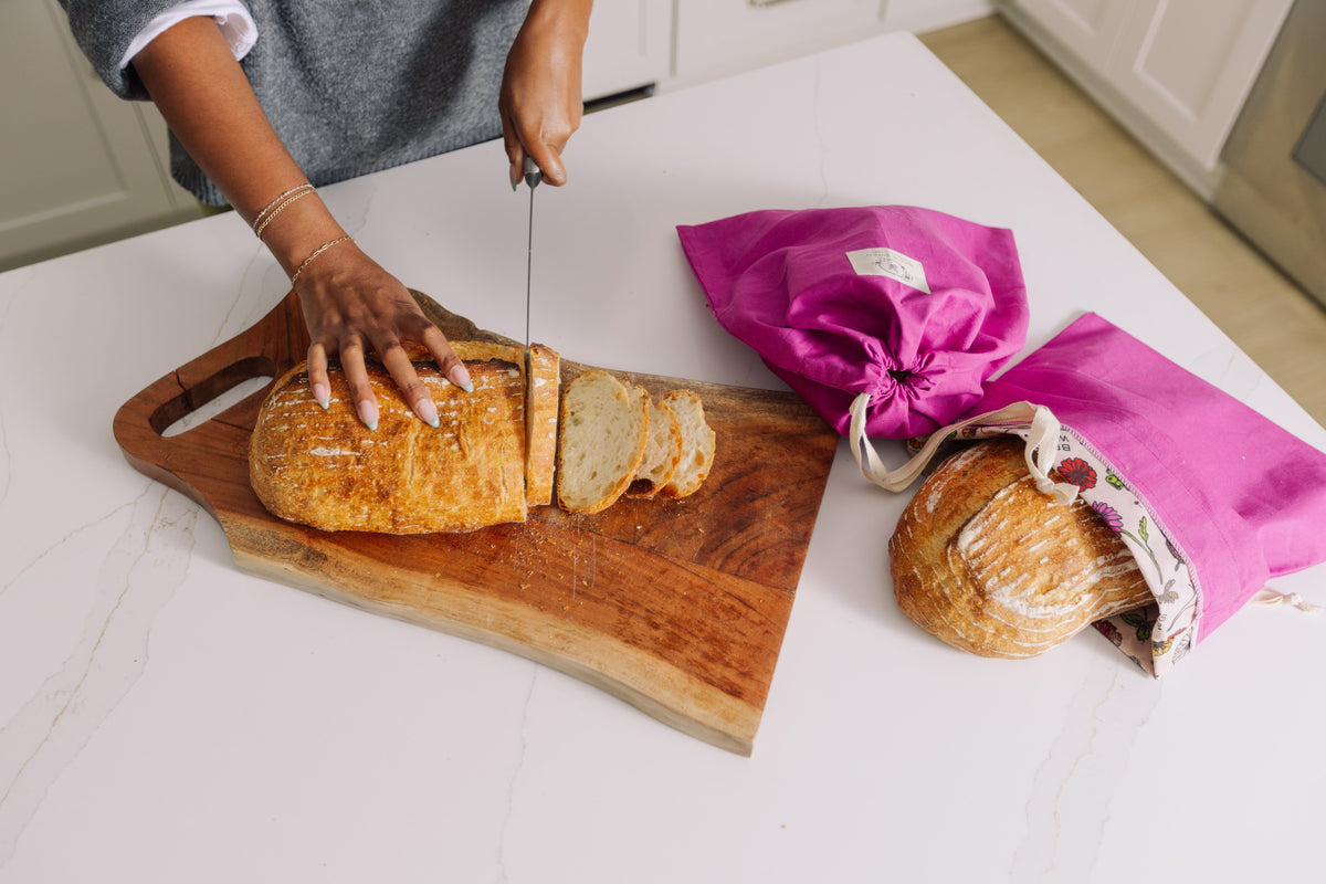 Person slicing bread on a wooden cutting board with Meadow Magic reusable bags in the background.