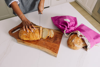 Person slicing bread on a wooden cutting board with Meadow Magic reusable bags in the background.