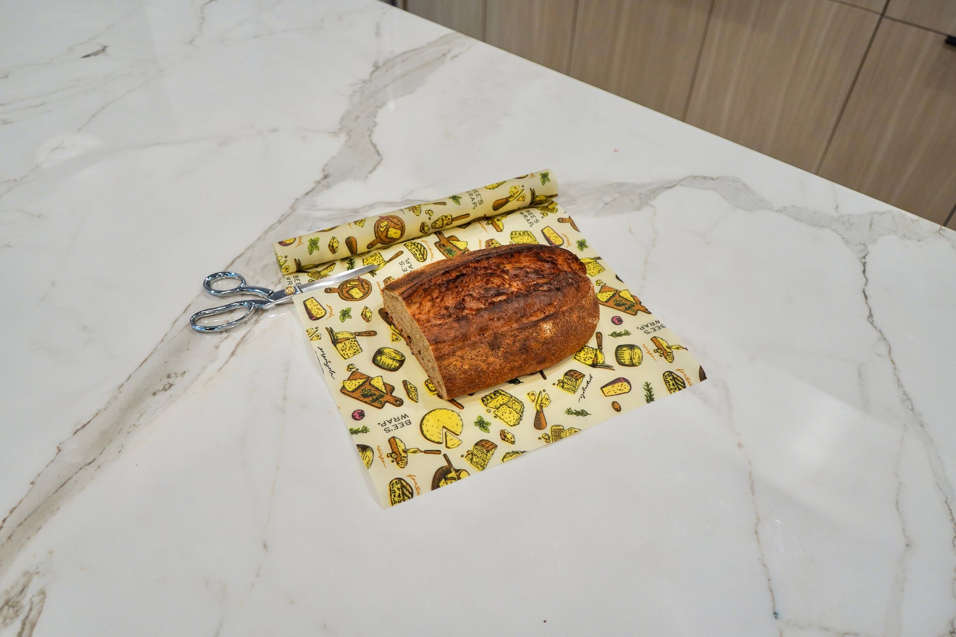 A loaf of bread placed on a reusable Bees Wrap Roll with the Wedges n Wheels print and blue-handled scissors on a white marble kitchen countertop.